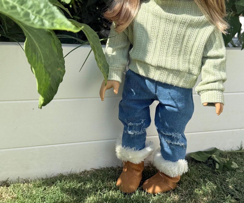 Puede incluir: Una mu&ntilde;eca con un su&eacute;ter verde claro y jeans azules con detalles desgastados. La mu&ntilde;eca lleva botas de piel sint&eacute;tica marr&oacute;n.