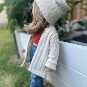 Op de afbeelding: Een pop die een witte kabelgebreide cardigan, een rood tanktop, blauwe jeans en een witte kabelgebreide beanie draagt.