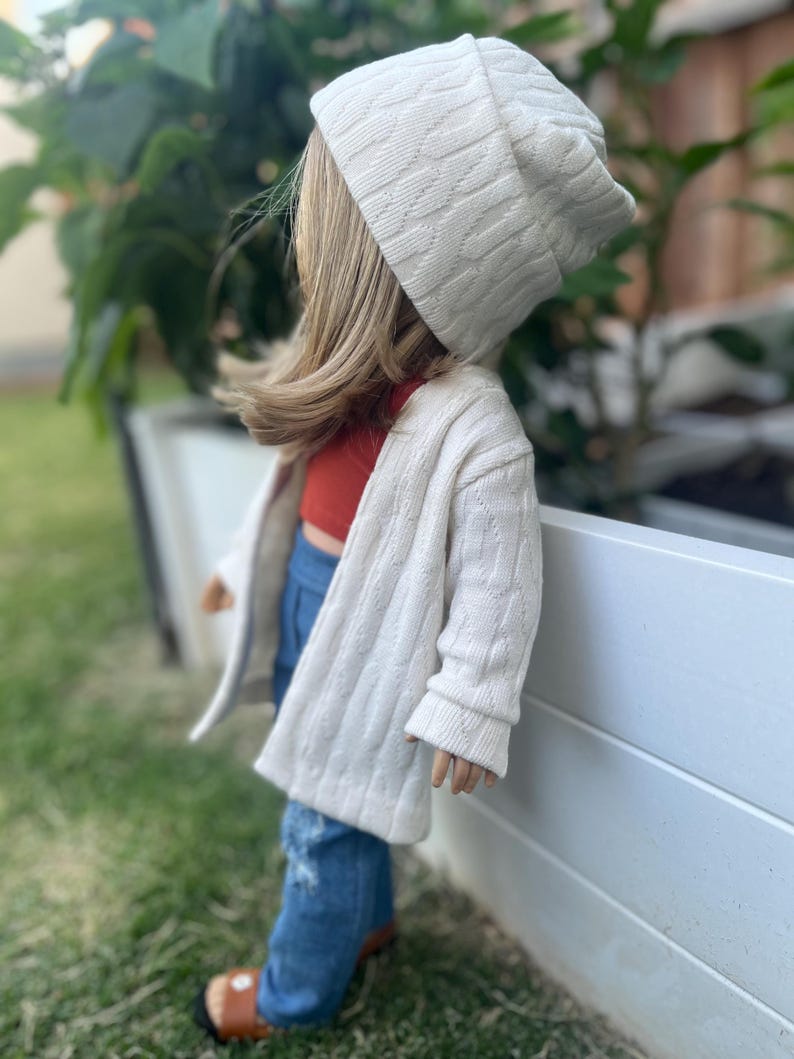 Op de afbeelding: Een pop die een witte kabelgebreide cardigan, een rood tanktop, blauwe jeans en een witte kabelgebreide beanie draagt.