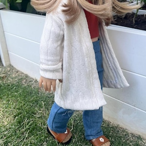 Op de afbeelding: Een pop die een witte cardigan, blauwe jeans en bruine sandalen draagt. De pop heeft lang blond haar en staat op een grasveld.