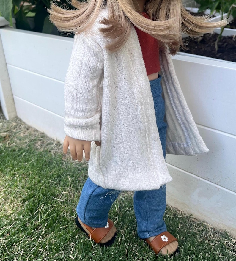 Op de afbeelding: Een pop die een witte cardigan, blauwe jeans en bruine sandalen draagt. De pop heeft lang blond haar en staat op een grasveld.