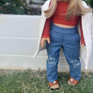 Op de afbeelding: Een pop die een rood topje draagt, een blauwe jeans met distressed details en een witte cardigan. De pop heeft lang blond haar en draagt bruine sandalen.