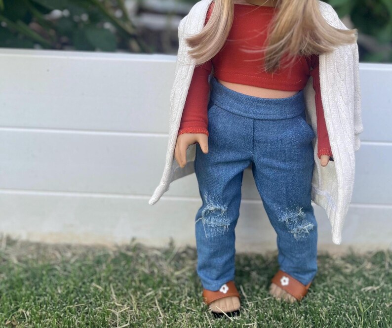 Op de afbeelding: Een pop die een rood topje draagt, een blauwe jeans met distressed details en een witte cardigan. De pop heeft lang blond haar en draagt bruine sandalen.