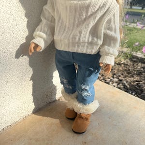 Puede incluir: Una mu&ntilde;eca con un su&eacute;ter blanco, pantalones vaqueros azules y botas marrones con ribete de piel blanca.