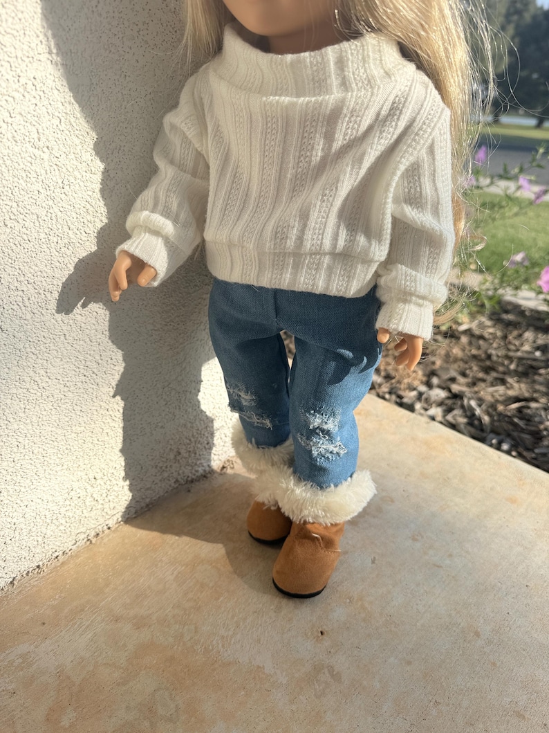 Puede incluir: Una mu&ntilde;eca con un su&eacute;ter blanco, pantalones vaqueros azules y botas marrones con ribete de piel blanca.
