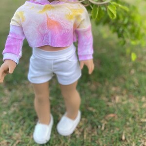 K&ouml;nnte beinhalten: Ein tie-dye-farbener, verk&uuml;rzter Hoodie mit einem wei&szlig;en Bund und wei&szlig;en Shorts. Der Hoodie ist pink, gelb und orange. Die Shorts sind wei&szlig; und haben einen Bund.