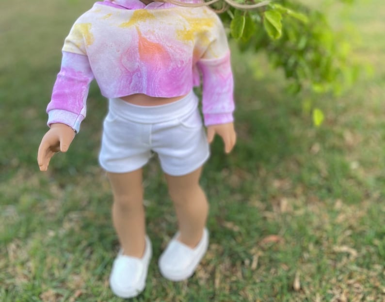 K&ouml;nnte beinhalten: Ein tie-dye-farbener, verk&uuml;rzter Hoodie mit einem wei&szlig;en Bund und wei&szlig;en Shorts. Der Hoodie ist pink, gelb und orange. Die Shorts sind wei&szlig; und haben einen Bund.