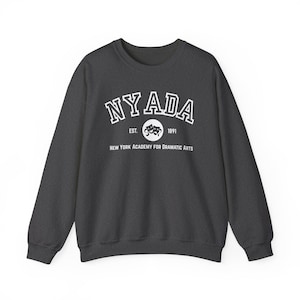 May include: A dark gray sweatshirt with the text "NYADA" in white, arched across the top. Below, a circular emblem with theater masks and the text "EST. 1891". The words "NEW YORK ACADEMY FOR DRAMATIC ARTS" are printed below the emblem.