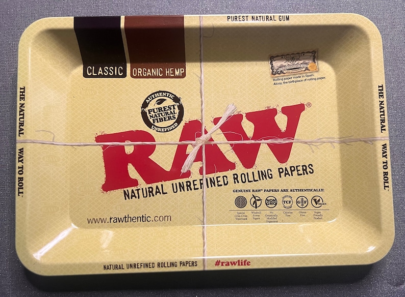以下が含まれることがあります： 赤と黒のRAWロゴがついた長方形の黄色の金属製のローリングトレイ。トレイは紐で結ばれており、「The Natural Way to Roll」と「Natural Unrefined Rolling Papers」の文字が書かれています。