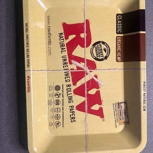 以下が含まれることがあります： 茶色の縁取りと黄色の背景を持つ金属製のローリングトレイ。トレイにはRAWのロゴと「The Natural Way to Roll」と「Classic Organic Hemp」の文字が書かれています。