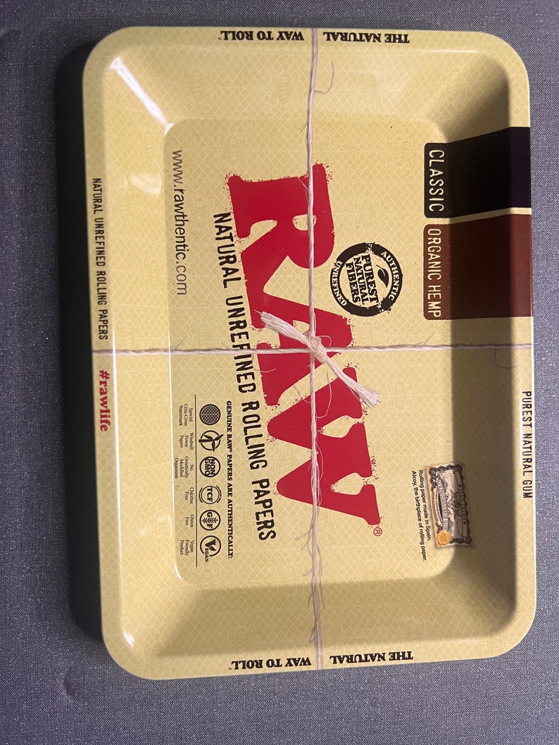 以下が含まれることがあります： 茶色の縁取りと黄色の背景を持つ金属製のローリングトレイ。トレイにはRAWのロゴと「The Natural Way to Roll」と「Classic Organic Hemp」の文字が書かれています。