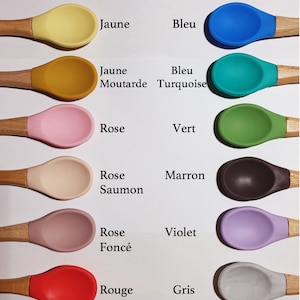 May include: A set of ten silicone baby spoons with wooden handles, each spoon is a different colour: yellow, mustard yellow, pink, peach, light brown, red, blue, turquoise, green, and purple. The spoons are arranged in two rows of five, with the text "Différents coloris" at the top.