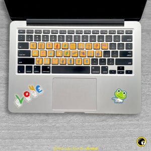 May include: A laptop keyboard with yellow sign language key stickers. The stickers are on the number and letter keys. There are also stickers that spell "LOVE" and a green frog sticker on the laptop. The laptop is on a gray surface.
