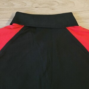 K&ouml;nnte beinhalten: Nahaufnahme eines schwarzen Poloshirts mit roter Schulterpartie und schwarzem Kragen. Das Shirt ist aus einem strukturierten Stoff gefertigt. Der Kragen ist gerippt.