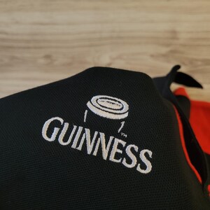 K&ouml;nnte beinhalten: Schwarzes Poloshirt mit dem Guinness-Logo, wei&szlig; gestickt. Das Logo zeigt den Namen Guinness und eine wei&szlig;e Grafik. Das Shirt hat einen roten Akzent an der Seite.