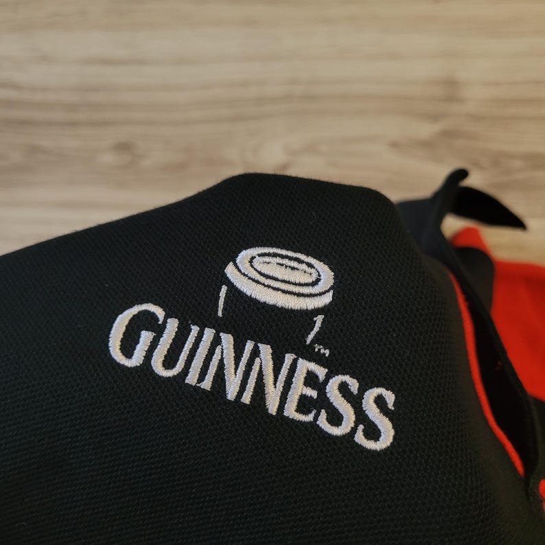 K&ouml;nnte beinhalten: Schwarzes Poloshirt mit dem Guinness-Logo, wei&szlig; gestickt. Das Logo zeigt den Namen Guinness und eine wei&szlig;e Grafik. Das Shirt hat einen roten Akzent an der Seite.