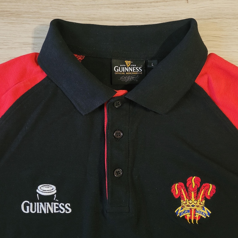 K&ouml;nnte beinhalten: Schwarzes Poloshirt mit roten Schulterpartien. Mit dem Guinness-Logo und einem Wappen mit Krone und Federn. Das Shirt hat einen Button-Down-Kragen und ist mit "Official Merchandise" gekennzeichnet.