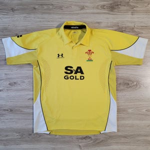 May include: Yellow rugby jersey with white and black accents. The jersey features the Under Armour logo, the text "SA GOLD", and a Welsh emblem. The collar has the text "HIRAETH".