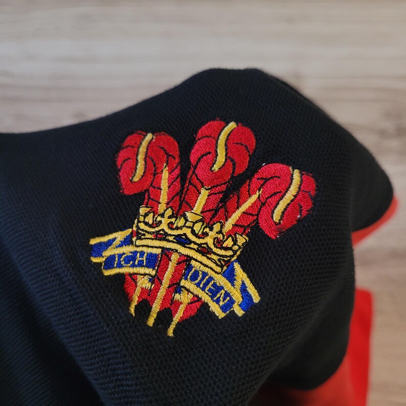 K&ouml;nnte beinhalten: Schwarzes Poloshirt mit rotem Besatz, mit einem gestickten Emblem einer Krone und drei roten und gelben Federn. Das Emblem enth&auml;lt den Text "ICH DIEN" in Blau und Gelb.