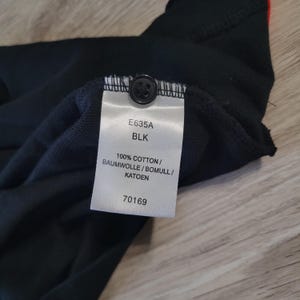 K&ouml;nnte beinhalten: Nahaufnahme eines schwarzen Kleidungsst&uuml;cketiketts mit dem Text "E635A BLK 100% COTTON / BAUMWOLLE / BOMULL / KATOEN 70169". Das Etikett ist an einem schwarzen Stoff mit einem Knopf befestigt.