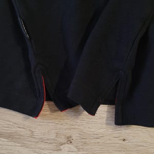 K&ouml;nnte beinhalten: Nahaufnahme eines marineblauen Poloshirts. Das Shirt hat einen strukturierten Stoff und rote Zierleisten am unteren Saum und an den Seitenschlitzen. Das Shirt ist gefaltet und zeigt die Details des Kragens und des Saums.
