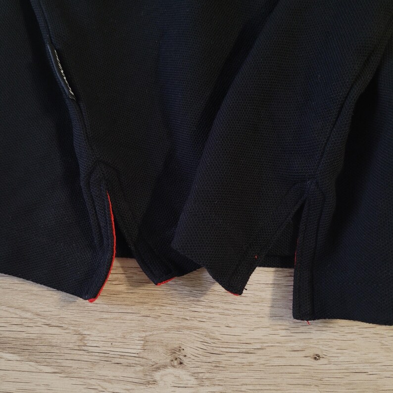K&ouml;nnte beinhalten: Nahaufnahme eines marineblauen Poloshirts. Das Shirt hat einen strukturierten Stoff und rote Zierleisten am unteren Saum und an den Seitenschlitzen. Das Shirt ist gefaltet und zeigt die Details des Kragens und des Saums.