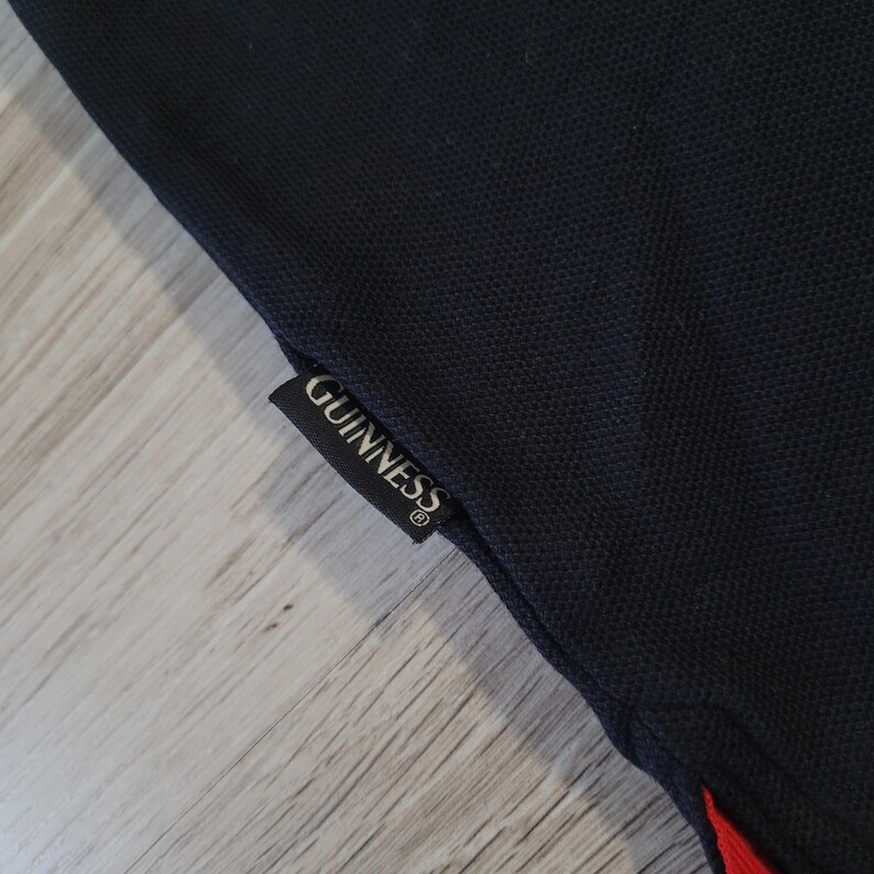 K&ouml;nnte beinhalten: Nahaufnahme eines marineblauen Poloshirts mit rotem Akzent. Das Shirt hat ein schwarzes Etikett mit dem wei&szlig;en Aufdruck "GUINNESS".