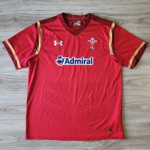May include: Red athletic jersey with a V-neck and short sleeves. The jersey features the Under Armour logo, the word "Admiral", and the Welsh Rugby Union emblem. Gold accents are on the sleeves and shoulders.
