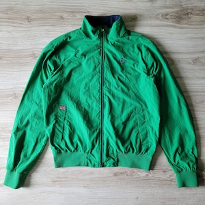 May include: Bright green zip-up jacket with a navy blue collar and cuffs. Features a small embroidered emblem and a Union Jack patch. The jacket has a ribbed hem and cuffs. The zipper is navy blue.