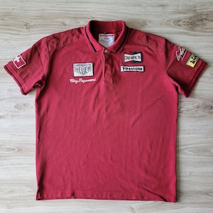 Könnte beinhalten: Rotes, kurzärmeliges Poloshirt mit schwarzem Besatz und verschiedenen gestickten Aufnähern, darunter "Champion Firestone", "Chronomatic Heuer Clay Regazzoni" und eine Schweizer Flagge.