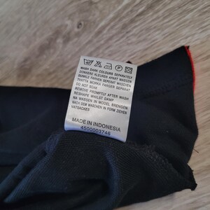 K&ouml;nnte beinhalten: Ein schwarzes Kleidungsst&uuml;cketikett mit Waschanweisungen in mehreren Sprachen. Das Etikett enth&auml;lt Symbole f&uuml;r Waschen, Trocknen und B&uuml;geln. Der Text auf dem Etikett enth&auml;lt "Wash dark colours separately" und "Made in Indonesia".