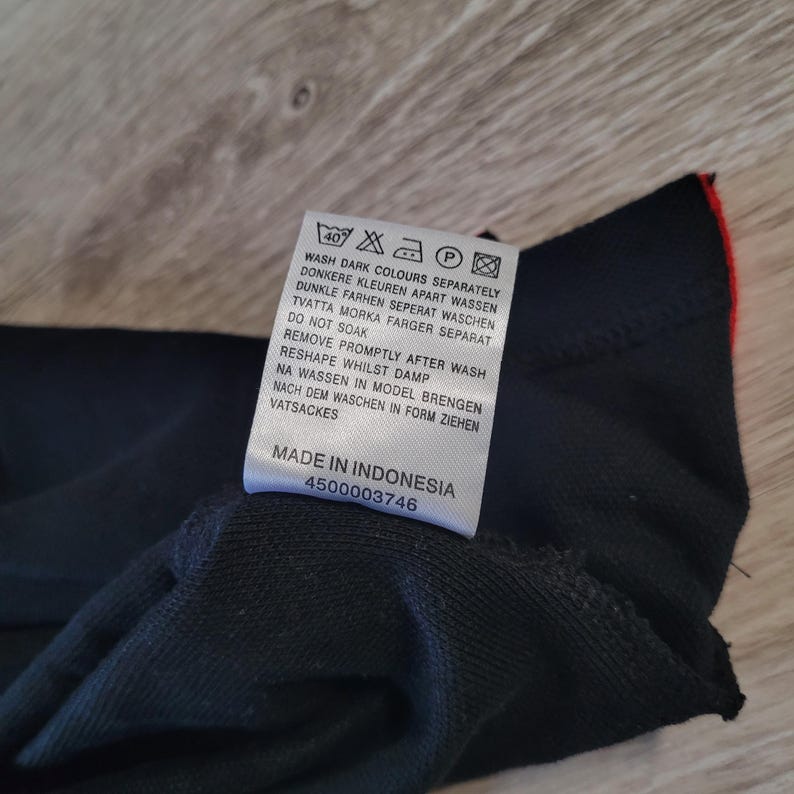 K&ouml;nnte beinhalten: Ein schwarzes Kleidungsst&uuml;cketikett mit Waschanweisungen in mehreren Sprachen. Das Etikett enth&auml;lt Symbole f&uuml;r Waschen, Trocknen und B&uuml;geln. Der Text auf dem Etikett enth&auml;lt "Wash dark colours separately" und "Made in Indonesia".