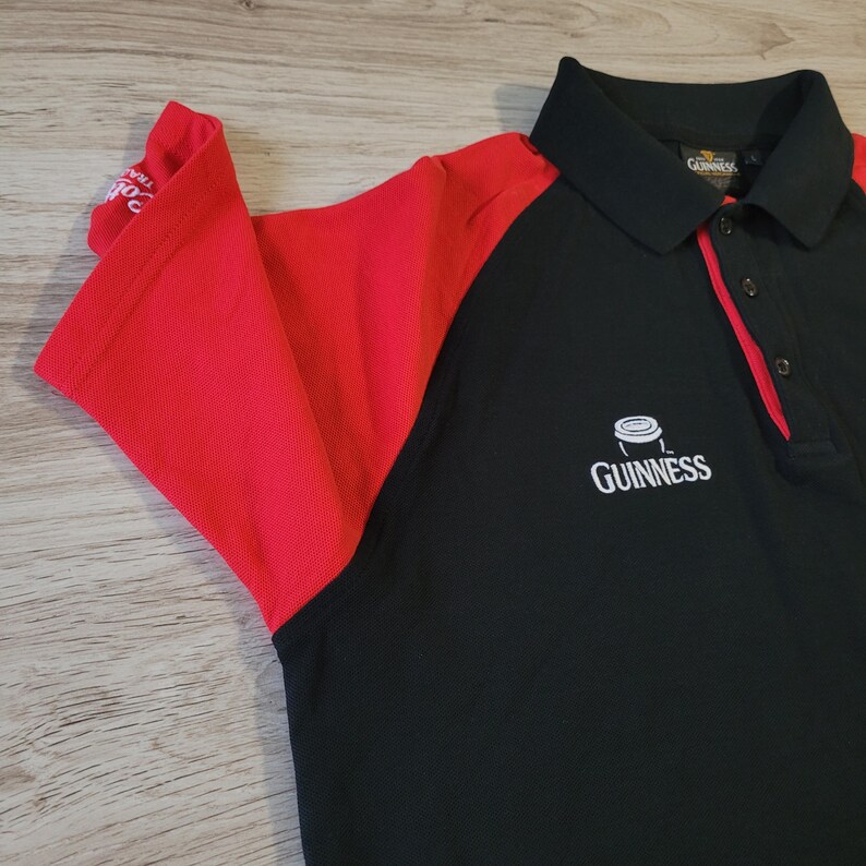 K&ouml;nnte beinhalten: Schwarzes Poloshirt mit roten &Auml;rmeln und Akzenten. Das Shirt hat das Guinness-Logo auf der Brust und einen schwarzen Kragen. Die &Auml;rmel sind leuchtend rot und das Shirt hat ein klassisches Polo-Design.