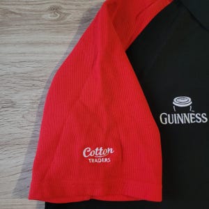 K&ouml;nnte beinhalten: Schwarz-rotes Poloshirt mit dem Guinness-Logo und dem Schriftzug "Cotton Traders". Das Shirt hat ein klassisches Design mit Kragen und kurzen &Auml;rmeln, ideal f&uuml;r Freizeitkleidung.