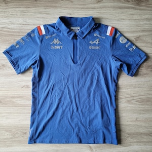 Puede incluir: Polo azul de manga corta con cuello con cremallera. La camiseta presenta logotipos de patrocinadores y el número 4. Rayas blancas y rojas en los hombros. La camiseta está hecha de un material transpirable.