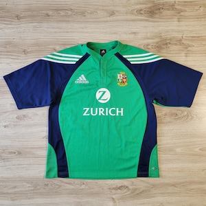 May include: Green and navy rugby jersey with white stripes on the shoulders. The jersey features the Adidas logo, a crest, and the word "ZURICH" in white. The shirt has short sleeves and a button-up collar.