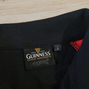 K&ouml;nnte beinhalten: Schwarzes Poloshirt mit schwarzem Kragen und roten Akzenten. Das Shirt zeigt ein Guinness Official Merchandise-Etikett mit dem Guinness-Logo und dem Text "ESTD 1759". Ein Gr&ouml;&szlig;enetikett "L" ist ebenfalls vorhanden.