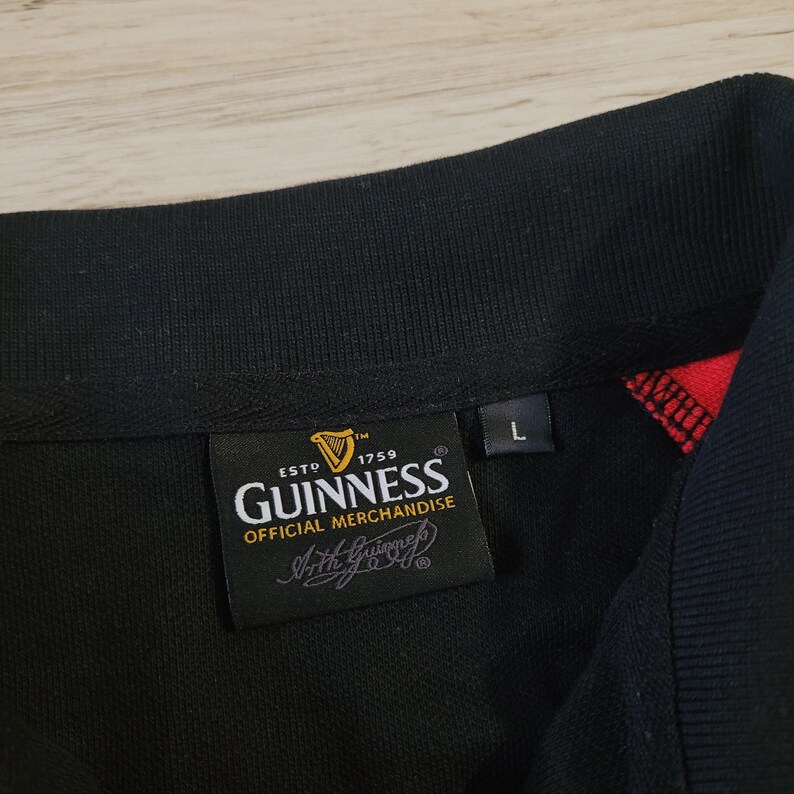 K&ouml;nnte beinhalten: Schwarzes Poloshirt mit schwarzem Kragen und roten Akzenten. Das Shirt zeigt ein Guinness Official Merchandise-Etikett mit dem Guinness-Logo und dem Text "ESTD 1759". Ein Gr&ouml;&szlig;enetikett "L" ist ebenfalls vorhanden.