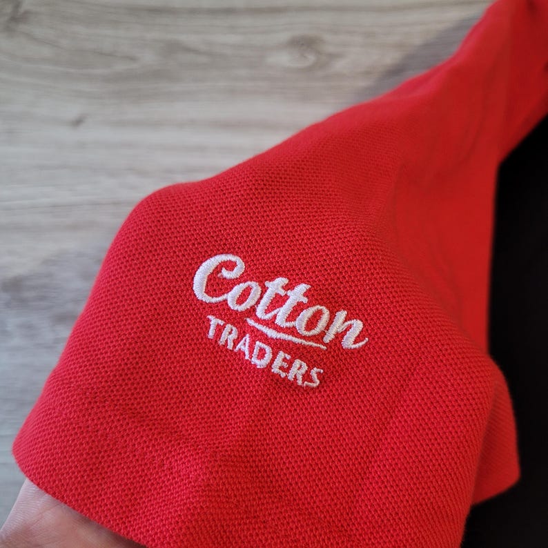 K&ouml;nnte beinhalten: Nahaufnahme eines roten Polo&auml;rmels mit dem gestickten Text "Cotton Traders" in Wei&szlig;. Der Stoff hat ein strukturiertes Aussehen. Das Shirt ist wahrscheinlich aus Baumwolle.