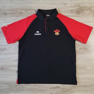 Può includere: Polo nero e rosso a maniche corte. La maglietta ha un corpo nero con maniche rosse e un colletto nero. Il logo Guinness è sul petto, insieme a un emblema gallese. La maglietta ha un colletto button-down.