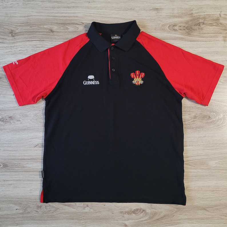 K&ouml;nnte beinhalten: Schwarzes und rotes Poloshirt mit kurzen &Auml;rmeln. Das Shirt hat einen schwarzen K&ouml;rper mit roten &Auml;rmeln und einen schwarzen Kragen. Das Guinness-Logo befindet sich auf der Brust, zusammen mit einem walisischen Emblem. Das Shirt hat einen Button-Down-Kragen.
