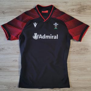 May include: Black and red short-sleeve jersey with a V-neck collar. The jersey features the word "Admiral" in white, along with a logo and the Welsh emblem. Red stripes adorn the sleeves.