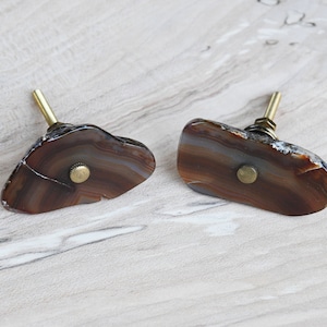 May include: Two agate drawer pulls with brass hardware. The agate is brown with white and gray banding. The pulls are in focus and the background is blurred.