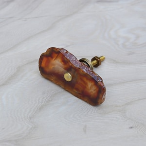 May include: A brown agate drawer pull with a gold metal knob and screw. The agate is a natural stone with a unique pattern.