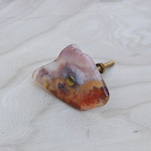 May include: A unique agate knob with a brass screw, featuring a mix of white, pink, and orange colors.