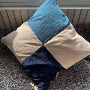 May include: Square corduroy cushion with a patchwork design. The cushion features alternating squares of beige, teal, and navy blue. The cushion is resting on a grey, textured surface.