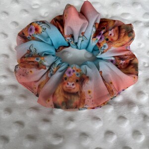 May include: A fabric scrunchie with a watercolor design featuring a Highland cow with a floral crown. The scrunchie has a blue and pink gradient background with floral accents. This hair accessory is a cute and stylish addition to any hairstyle.