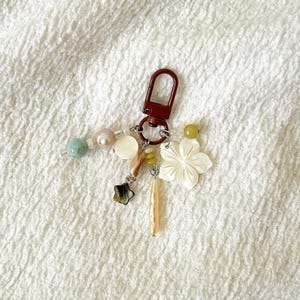 May include: A decorative keychain featuring a maroon clasp and an assortment of charms. The charms include a white flower, a star, and pastel-colored beads. The keychain is designed for attaching to keys or a bag.