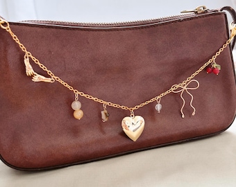 Puede incluir: Una cadena dorada con abalorios unidos a un bolso de cuero marrón. Los abalorios incluyen un corazón, un par de manos, una pequeña cuenta, un cristal y un pequeño lazo con bayas rojas.