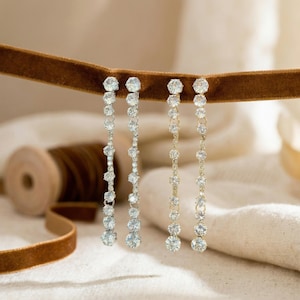 May include: Two pairs of dangling earrings with clear, round gemstones. One pair has a silver-colored setting, and the other has a gold-colored setting. The earrings are hanging from a brown velvet ribbon against a neutral background.